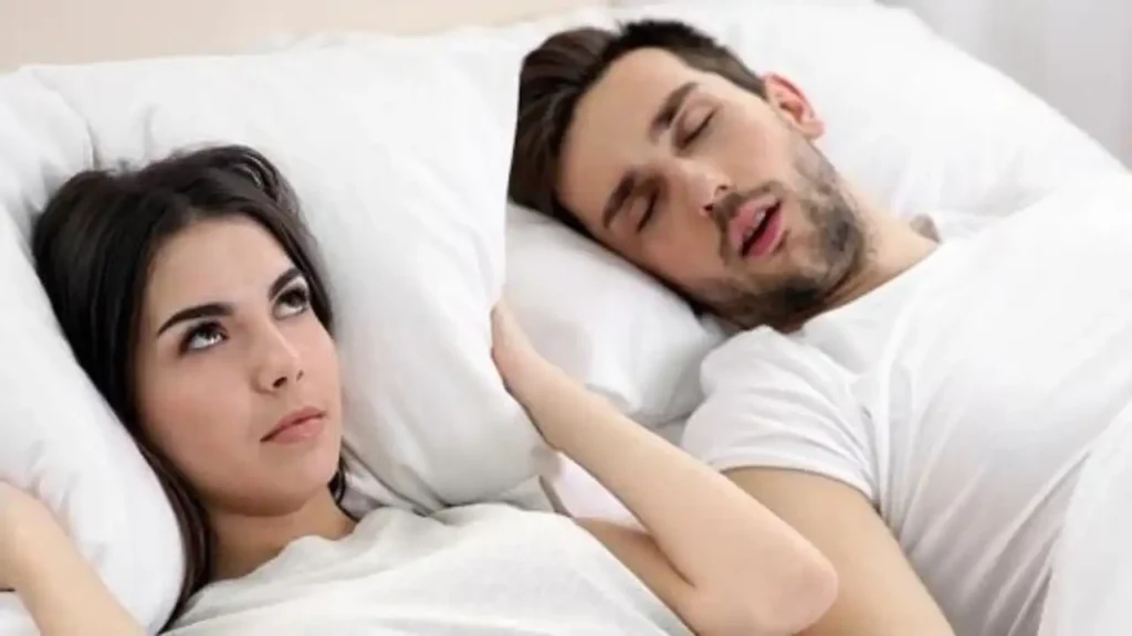 A woman lies in bed with her eyes open, looking annoyed, while a man next to her is in a deep sleep, his mouth open. Both are wearing white shirts and resting on white pillows.